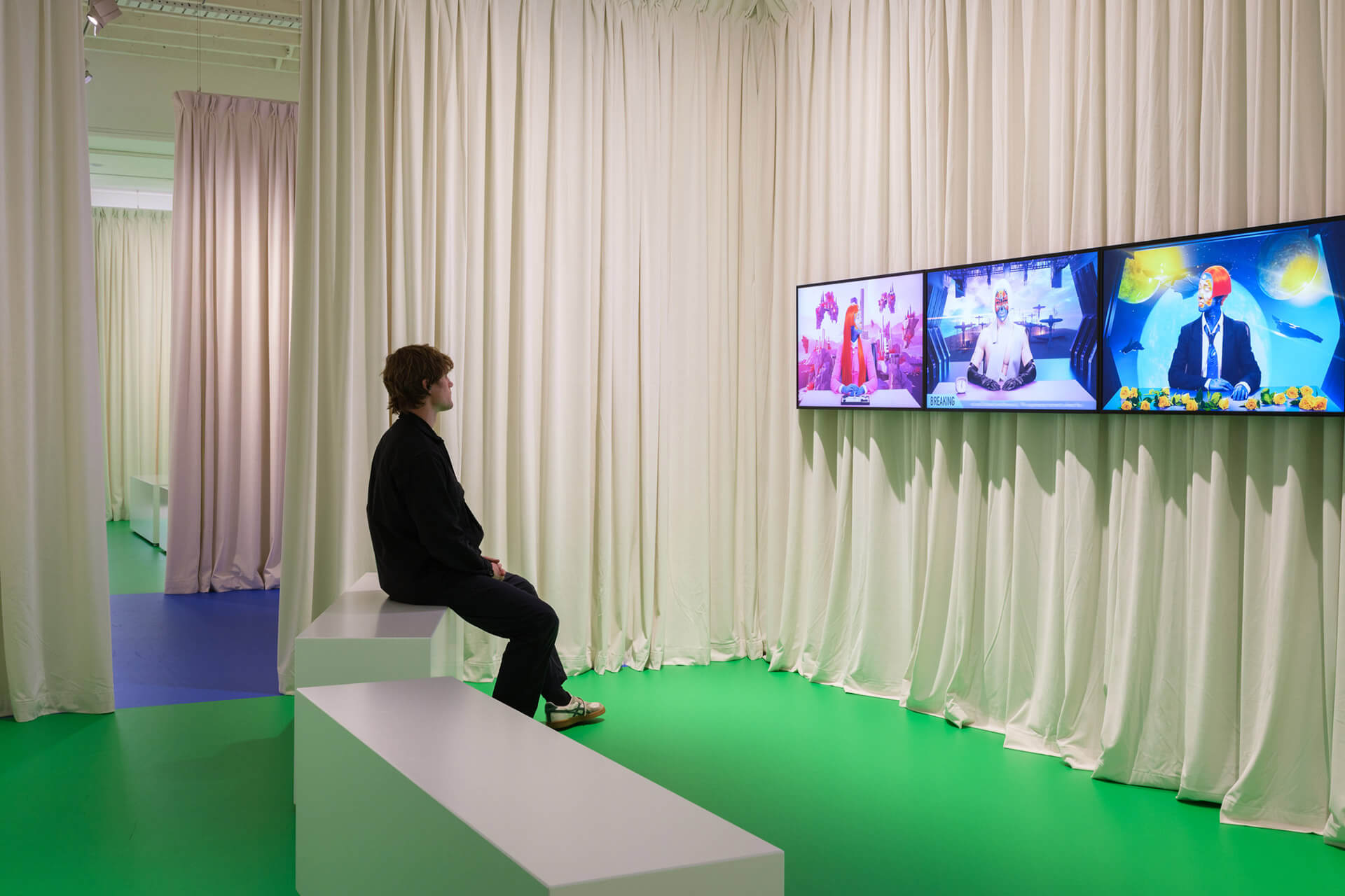 Visitor seated viewing Sin Wai Kin's "The Breaking Story " large screen video displaying colorful imagery with multiple figures in curtained gallery space