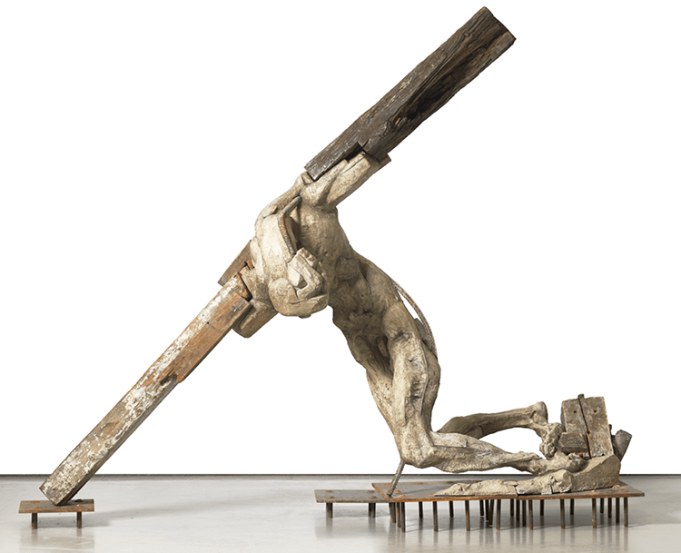 A large abstract sculpture made from fibreglass, rocks, wood, metal, resembling a figure with arms on a cross.