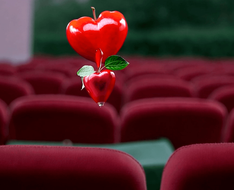 Still image of a video work, with an apple in the shape of a heart seemingly floating over red chairs in a theatre.