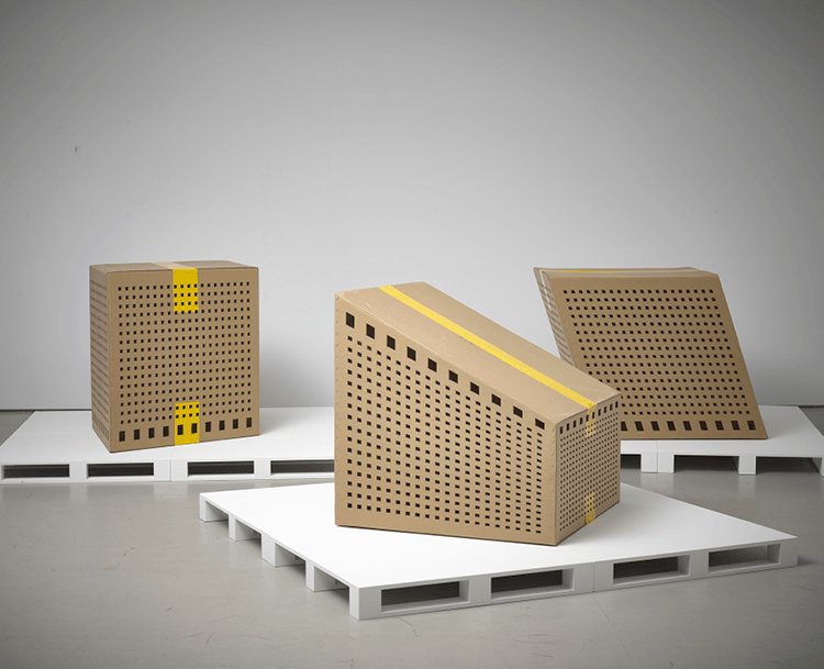 Three large installations of cubes that look like buildings made out of cardboard. They are each sitting on separate white pallet bases.